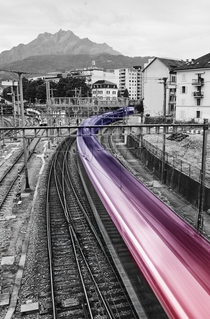 Vorbeirauschender Zug in Violett und Pink auf Schwarzweiss-Gleisen, im Hintergrund der Pilatus